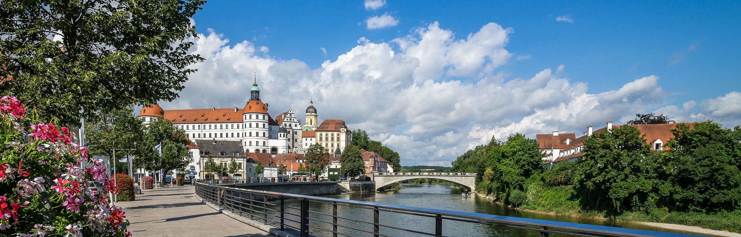 Fluss mit Brücke, rechts Bäume und Häuser, links Blumen, Baum und Schloss im Hintergrund bei blauem Himmel.