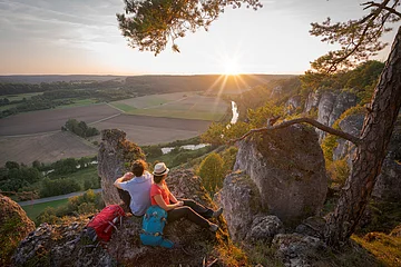 Zwei Wanderer sitzen auf Felsen mit Blick auf ein Tal und die untergehende Sonne am Horizont.