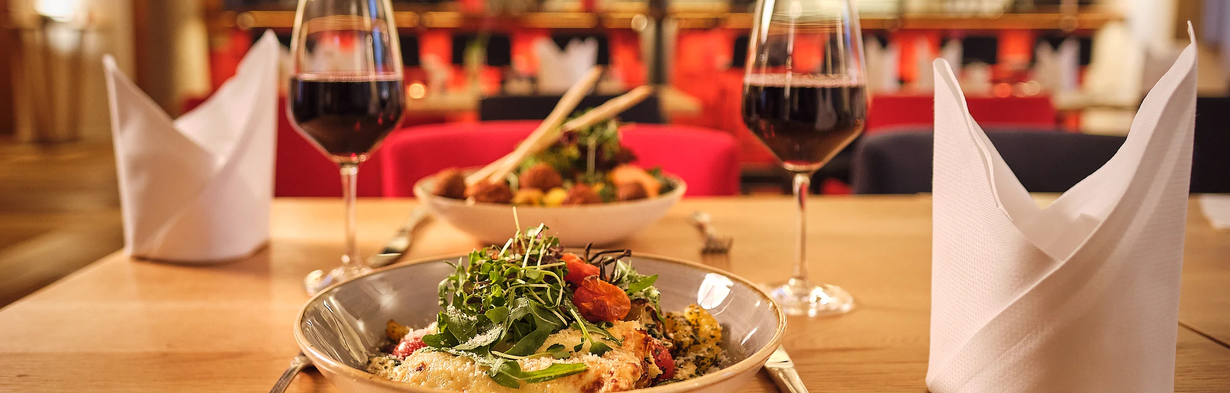 Tisch mit zwei Tellern, zwei Gläsern Rotwein, Besteck und zwei gefalteten weißen Servietten in einem Restaurant.