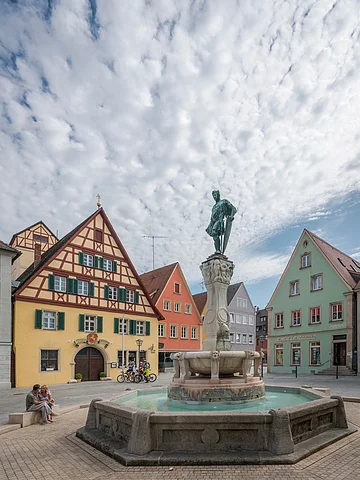 Brunnen mit Statue auf einem Platz, umgeben von Fachwerkhäusern und Menschen, unter bewölktem Himmel.