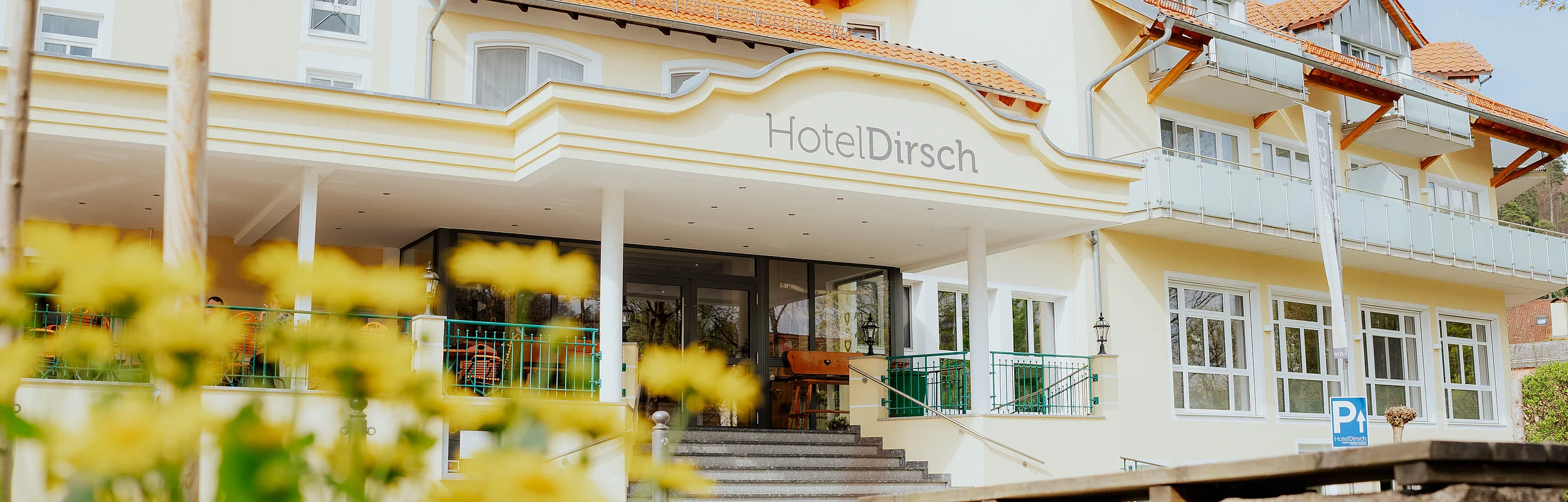 Eingangsbereich des Hotels Dirsch mit Stufen und gelben Blumen im Vordergrund bei Tageslicht.