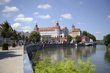 Flussufer mit Promenade und Bäumen, im Hintergrund Schloss mit Türmen und Brücke bei klarem Himmel.