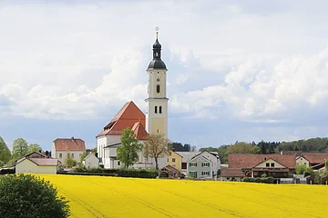 Dorf mit Kirche, Häusern und gelbem Rapsfeld unter bewölktem Himmel.