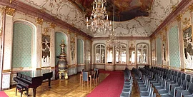 Prunkvoller Saal mit Kronleuchtern, roten Teppich, Klavier, Stühlen und goldverzierten Wänden und Decke.