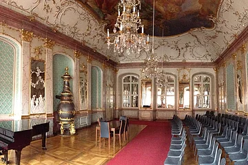 Prunkvoller Saal mit Kronleuchtern, roten Teppich, Klavier, Stühlen und goldverzierten Wänden und Decke.