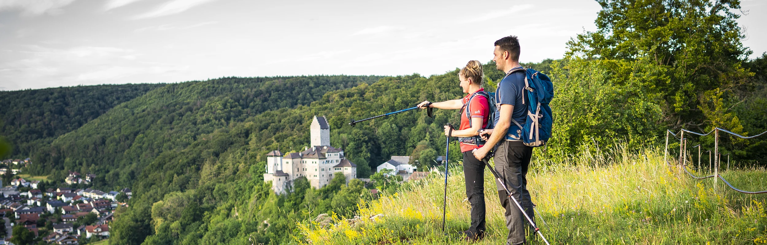 Ein Frau und ein Mann beide in Wanderkleidung und mit Rucksäcken stehen auf dem grün bewachsenem Michelsberg und schauen auf Kipfenberg hinunter. Im Hintergrund ist die Burg Kipfenberg inmitten von grünem Wald etwas zu erkennen. Der Himmel ist leicht grau und bewölkt.