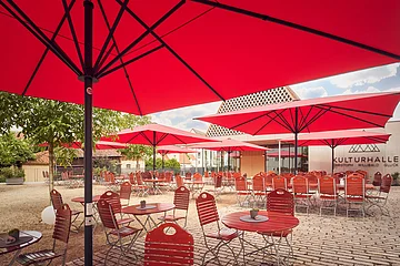 Außenbereich eines Cafés mit roten Sonnenschirmen und roten Stühlen vor einem Gebäude mit Schriftzug Kulturhalle.