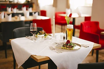 Tisch in Restaurant mit weißer Tischdecke, Kerze, Glas Rotwein, Glas Bier und angerichtetem Essen.