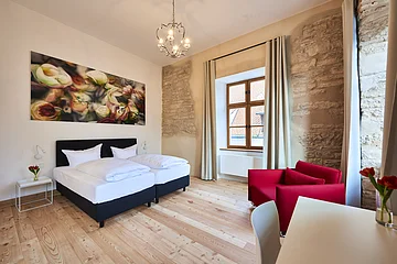 Hotelzimmer mit Doppelbett, rotem Sessel, Holzboden, Fenster mit Vorhängen und Wandbild mit Blumenmotiv.