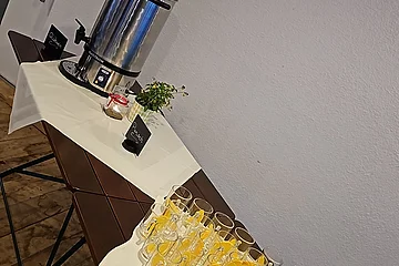 Tisch mit Gläsern und Orangenscheiben, Thermoskanne, Blumen und Kerze vor weißer Wand in Raum mit Holzboden.