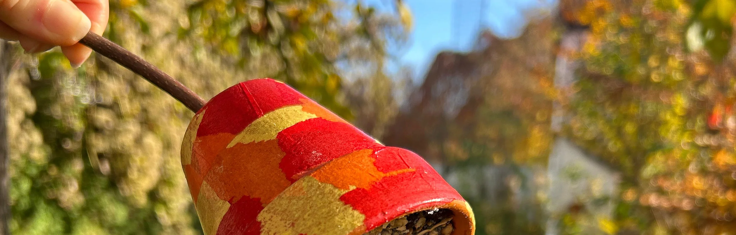 Hand hält bemalten Blumentopf mit Vogelfutter an einem Ast vor unscharfem Gartenhintergrund im Herbst