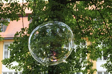 Eine riesige Seifenblase schwebt in der Höhe, hinter ihr die grünen Äste eines Baumes. Durch diese ist ein Gebäude zu erkennen.