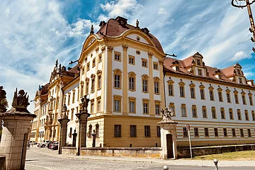 Zentrales Element des Bildes ist die Schlossresidenz Ellingen mit kräftigen Farben vor einem blauen Himmel. Links ist ein Pfeiler mit einer Statue zu sehen