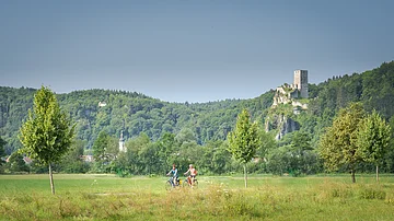 Zwei Radfahrer auf einem Weg vor grüner Wiese, Bäumen und bewaldetem Hügel mit Burgruine im Hintergrund.
