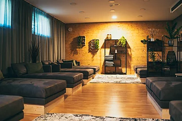 Raum mit mehreren gepolsterten Liegen, Holzwand, Regalen mit Pflanzen und Handtüchern, Vorhängen am Fenster.