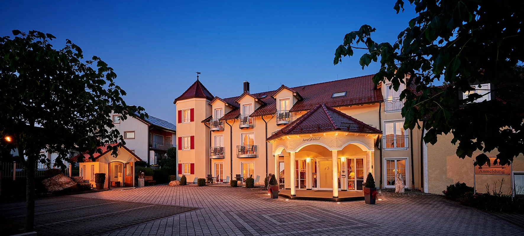 Beleuchtetes Hotelgebäude mit gepflastertem Vorplatz und Bäumen bei Abenddämmerung.