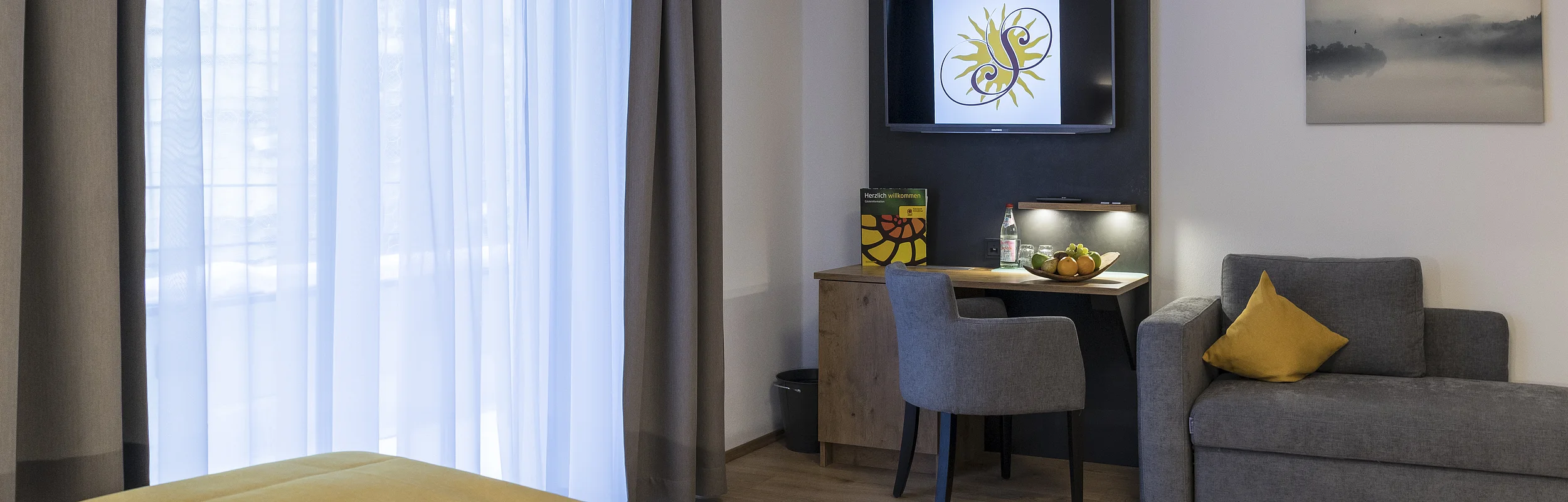 Hotelzimmer mit Bett, Schreibtisch, Fernseher, grauem Sessel und Sofa mit gelbem Kissen, helle Vorhänge am Fenster.