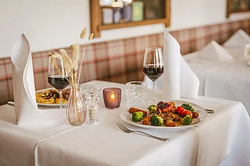 Tisch mit weißer Tischdecke, Teller mit Essen, zwei Weingläser, Vase mit Trockenblumen und Kerze in Restaurant.