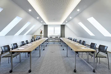 Konferenzraum mit zwei langen Tischen, Stühlen und Dachfenstern in einem hellen Raum mit Teppichboden.