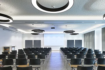 Konferenzraum mit Reihen schwarzer Stühle, Leinwand und runden Deckenleuchten in modernem Design.
