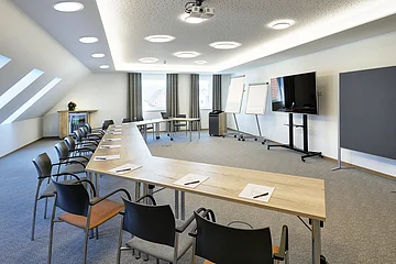 Konferenzraum mit U-förmigen Tischen, Stühlen, Flipcharts, großem Bildschirm und Dachfenstern.