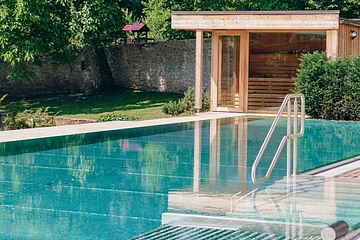 Außenpool mit Edelstahlgeländer und Holzhütte vor großer Steinmauer und Bäumen im Garten.