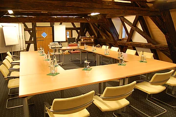 Konferenzraum mit U-förmigen Tischen, Stühlen, Wasserflaschen und Flipcharts unter Holzbalken.