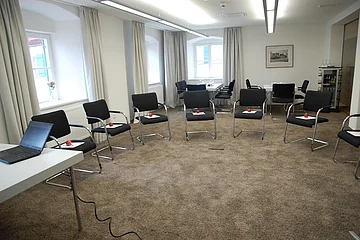 Konferenzraum mit Stühlen im Halbkreis, auf jedem Stuhl ein Apfel und ein Zettel, Laptop auf Tisch links.