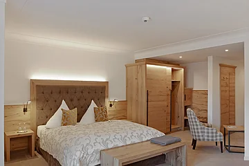 Hotelzimmer mit Doppelbett, Holzmöbeln, Sessel und Tisch in neutralen Farben und Beleuchtung.