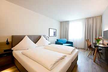 Hotelzimmer mit zwei Einzelbetten, blauem Sessel, Schreibtisch mit Stuhl und Fenster mit Vorhängen.