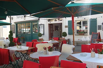 Außenbereich eines Cafés mit roten Stuhlhussen, weißen Tischdecken und grünen Sonnenschirmen.