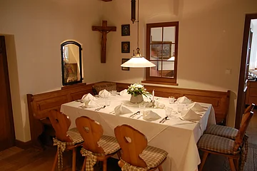 Esszimmer mit Holztisch, weißer Tischdecke, sechs Holzstühlen mit Kissen, Wandkreuz und Fenster mit Spiegel.