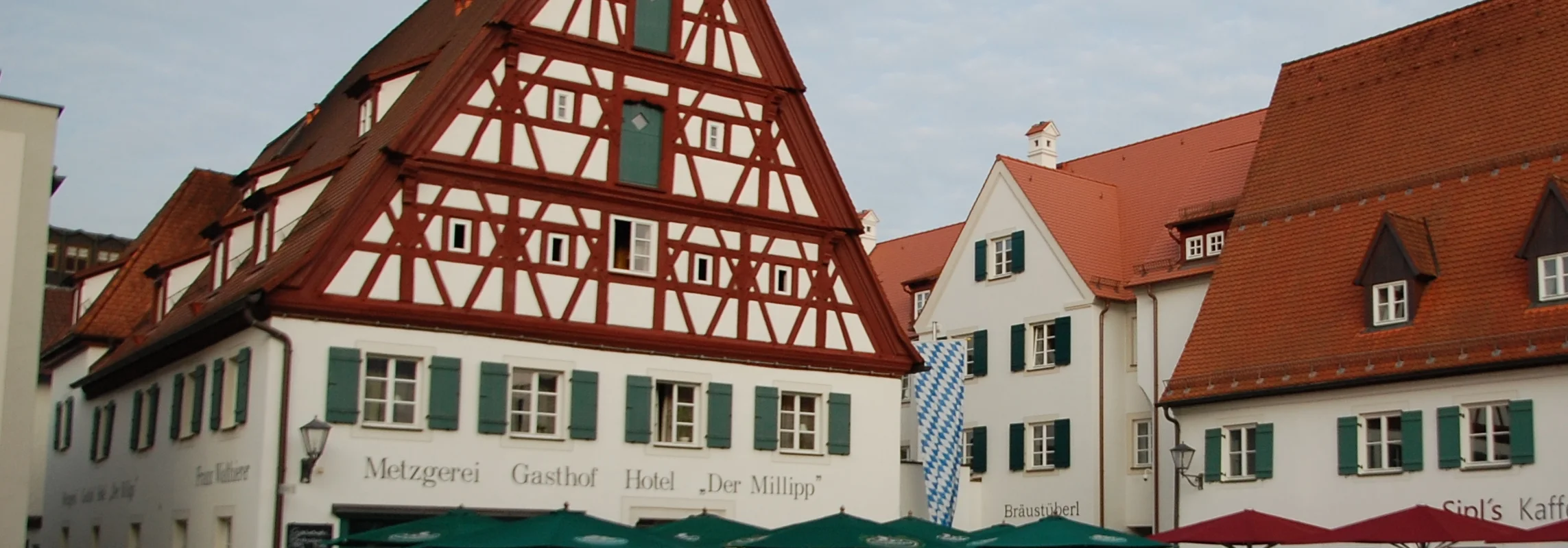 Traditionelle Fachwerkhäuser mit Außenbestuhlung und Sonnenschirmen an einem gepflasterten Platz.