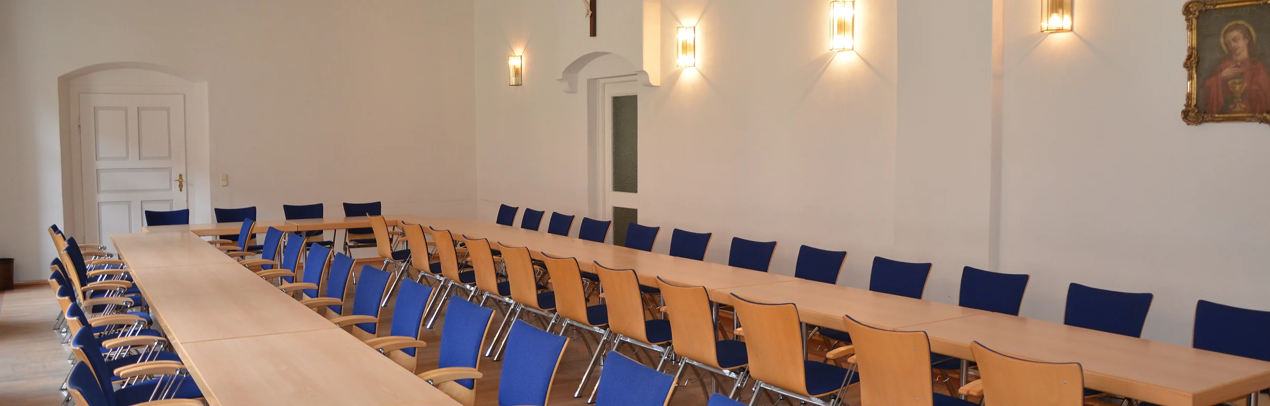 Konferenzraum mit langen Holztischen, blauen Stühlen, weißer Wand, Kreuz und Bild an der Wand, verziertem Deckenmuster