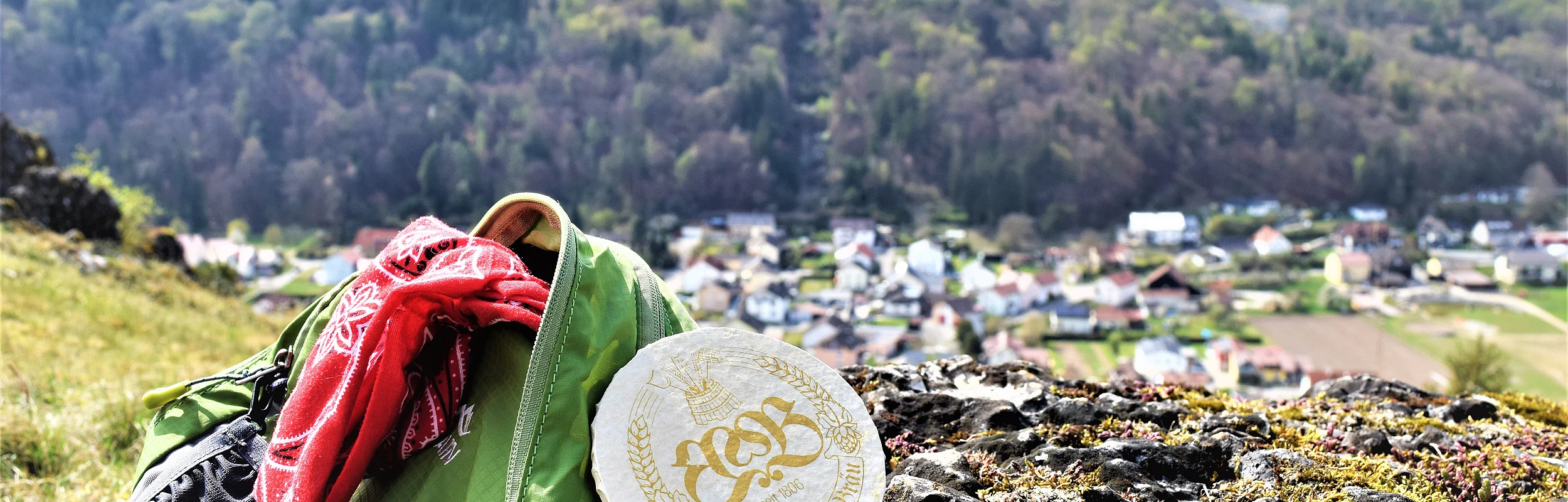 Grüner Rucksack mit rotem Tuch und rundem Bierdeckel auf Felsen, im Hintergrund Dorf und bewaldete Hügel.