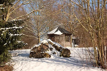 Schneebedeckter Wald mit kahlen Bäumen, einem großen Stein mit Schild und einer Holzhütte im Hintergrund.