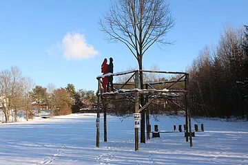 Holzplattform mit zwei Figuren, Schnee bedeckt, umgeben von Bäumen unter blauem Himmel