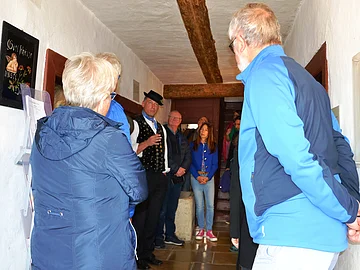 Gruppe von Menschen in einem engen Raum, einige hören einem Mann in traditioneller Kleidung zu.
