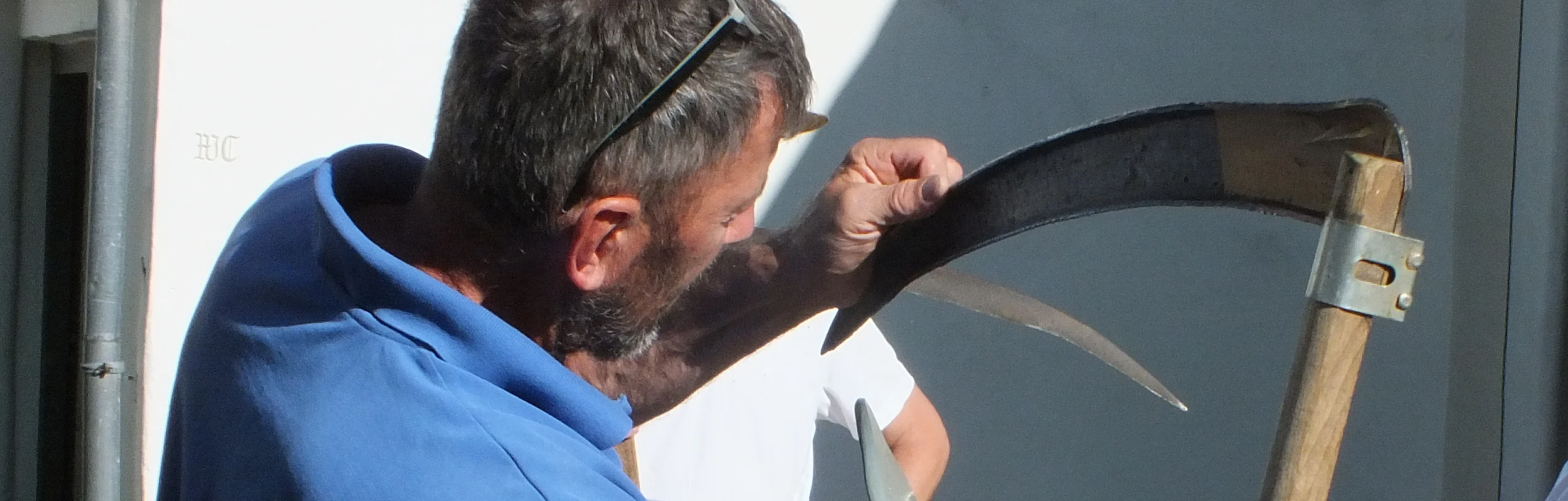 Zwei Männer schärfen eine Sense im Freien. Einer hält die Sense, der andere bearbeitet die Klinge.