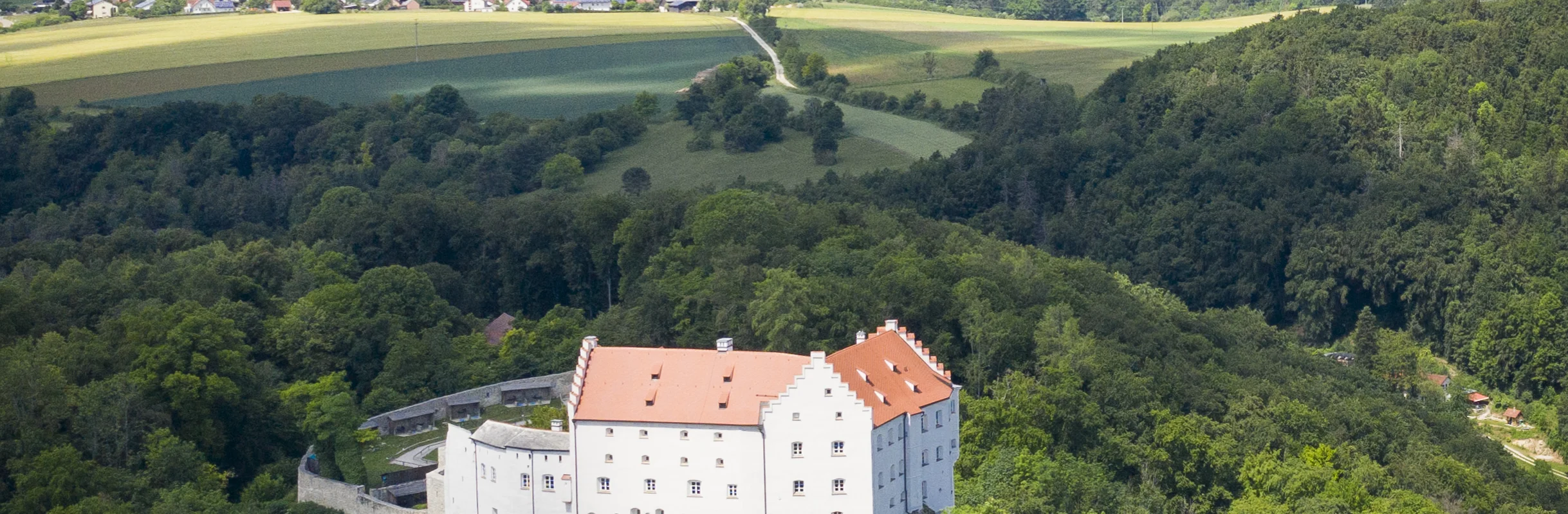 Luftaufnahme eines weißen Schlosses mit rotem Dach, umgeben von Wald und Feldern unter blauem Himmel.
