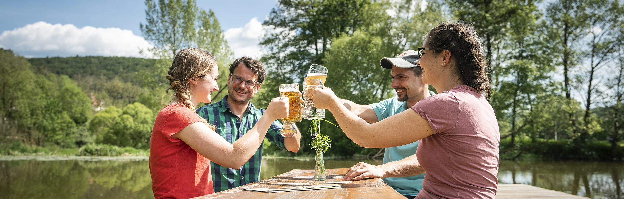 Vier Personen stoßen mit Biergläsern an einem Holztisch im Freien an, umgeben von Bäumen und einem See.