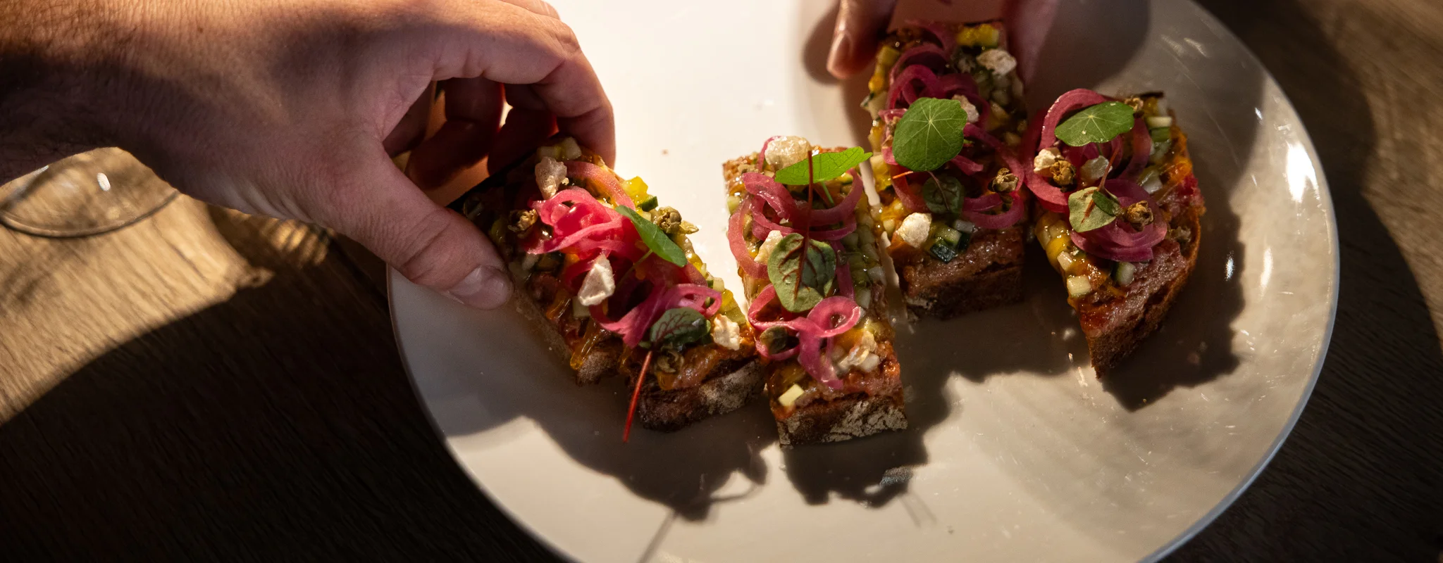 Zwei Hände nehmen kleine belegte Brotscheiben von einem weißen Teller auf einem Holztisch.