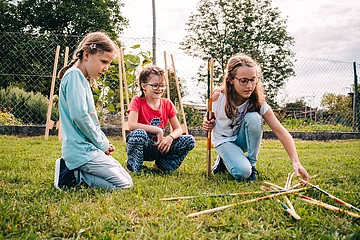 Drei Mädchen knien auf Gras und spielen mit langen Holzstäben im Garten vor einem Zaun.