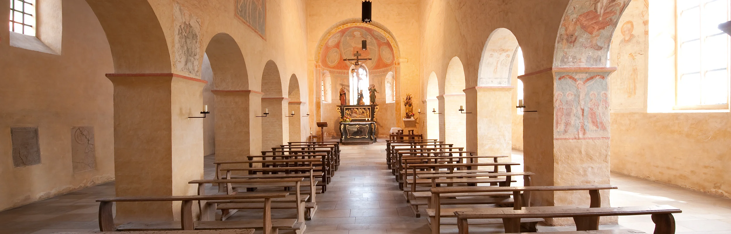 Innenraum einer Kirche mit Holzbänken, Säulen und Altar im Hintergrund, helle Wände mit Fresken und Fenstern