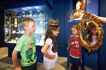 Drei Kinder sind im Kristallmuseum und ein Mädchen hat eine Krone auf und schaut in den Spiegel