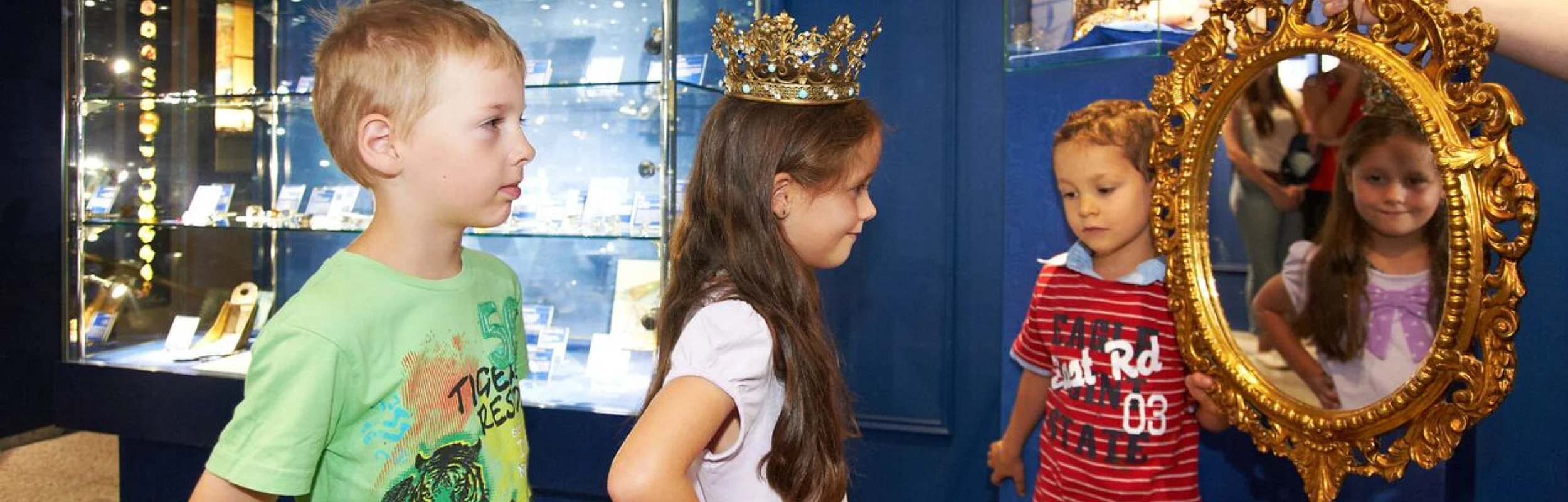 Drei Kinder sind im Kristallmuseum und ein Mädchen hat eine Krone auf und schaut in den Spiegel