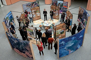 Menschen stehen in einem Kreis um Ausstellungstafeln mit Texten und Bildern in einer Halle.