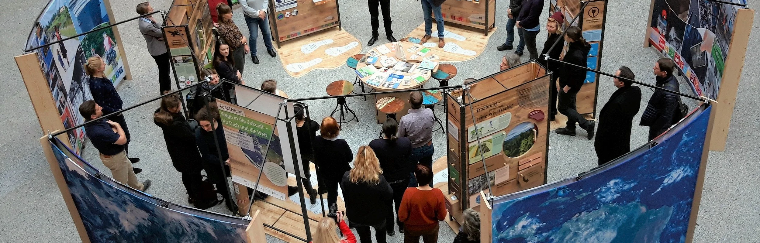 Menschen stehen in einem Kreis um Ausstellungstafeln mit Texten und Bildern in einer Halle.