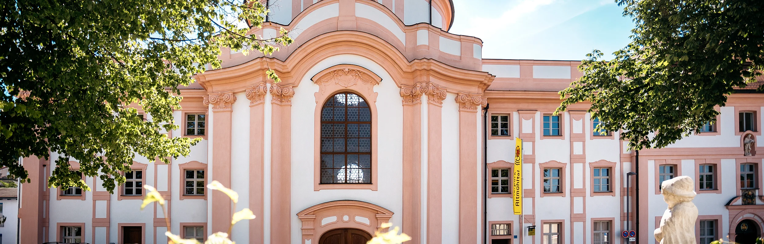 Barockgebäude mit großer Fensterfront, Statue auf dem Dach und Springbrunnen im Vordergrund bei Sonnenschein