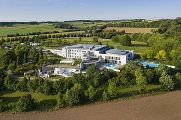 Luftaufnahme eines großen Gebäudekomplexes mit Schwimmbädern umgeben von Bäumen und Feldern.